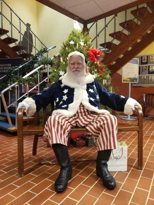 A man dressed as a Civil War era Santa Claus, wearing a blue jacket with white stars and red and white striped pants, sits on a bench in front of a Christmas tree.