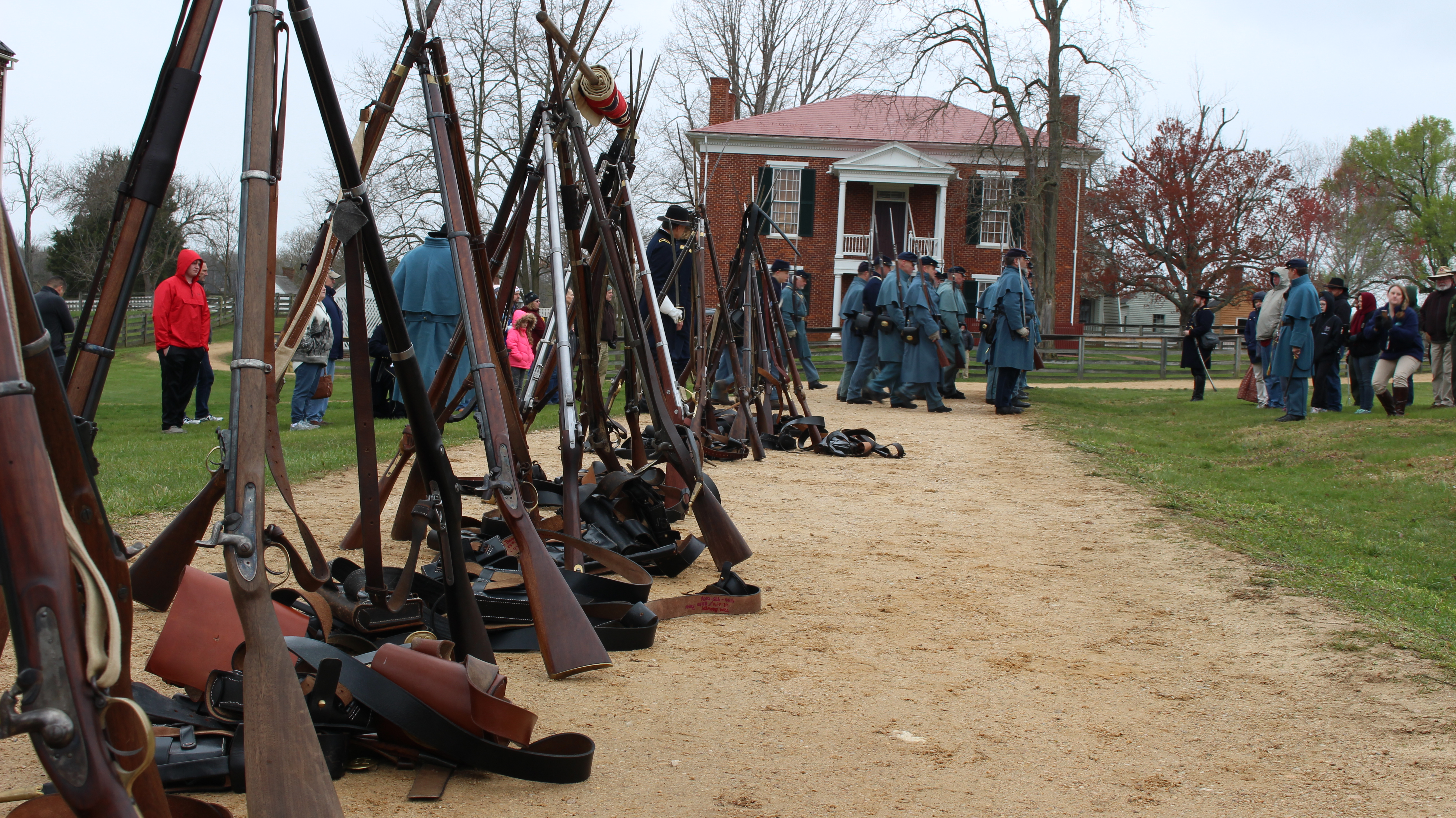 Three rifles leaned together to form a standing triangular prism, repeated down the length of a gravel road with two story brick courthouse at the end. Flags and accoutrements hang and lay beside the stacked arms.