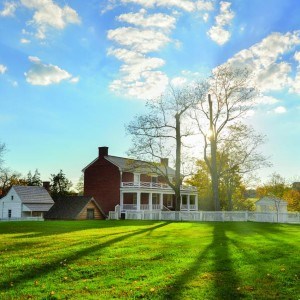 A two story brick house with white porches on both floors sits between a blanket of bright green grass and a deep blue sky. The sun peeks from behind a tree in front of the house and casts long shadows on the grass. Three wooden outbuildings are visible.
