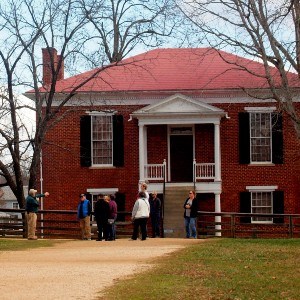 Two story brick courthouse building with dirt road leading to lower floor entrance. A prominent staircase leads to the second floor entrance with roofed landing and black door. Small group of visitors stand out front.