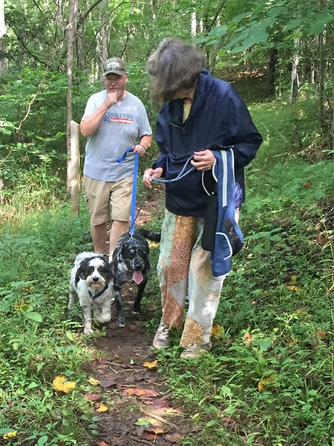 Two visitors are walking on a trail in the park with their two dogs.