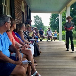 A row of visitors dressed in summer attire listen closely to the male park ranger presenting a program, all seated in a row on the historic Tavern's wooden porch.