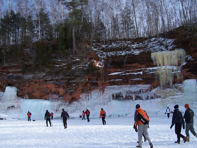 A large number of people walk across a snow-covered, frozen lake toward tall cliffs with ice formations.
