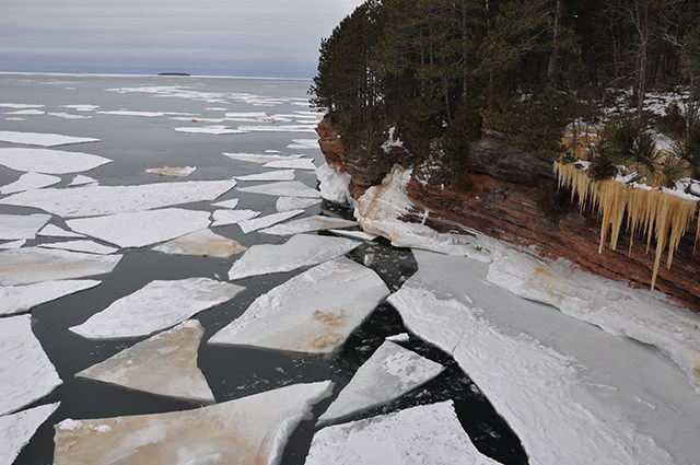 A rocky cliff covered in ice and snow near a lake with large chunks of ice floating.