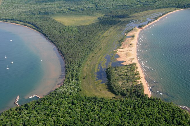 An aerial photo of a wooded narrow spot of an island, with water on both sides and a beach on the right.