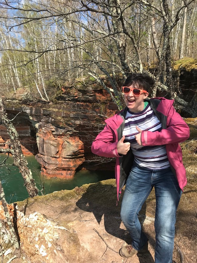 A hiker smiles and gives a thumbs up at a cliff overlooking a lake.