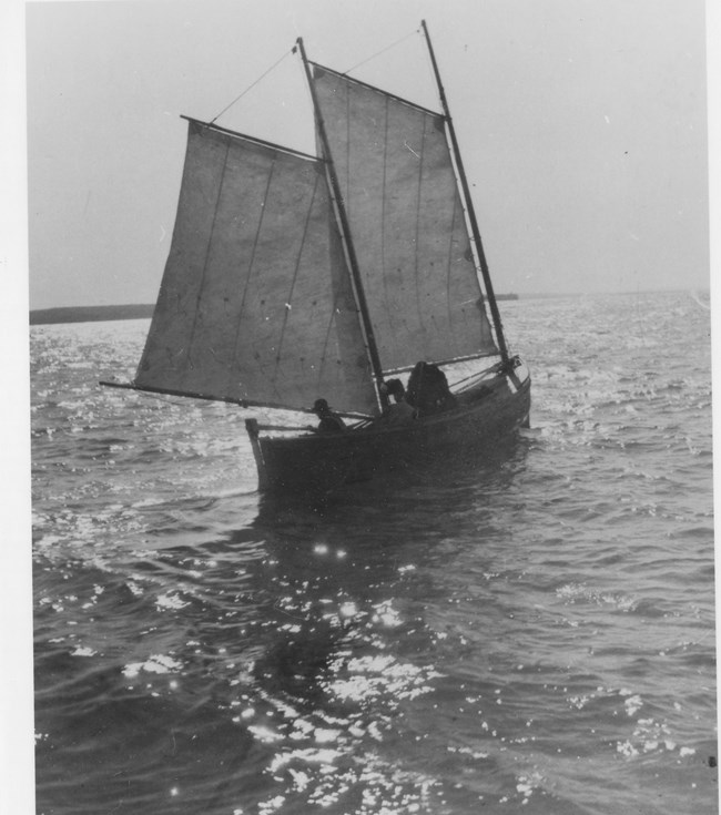 A black and white photo of a small sailboat with two sails up from mid 1900s on a large lake.