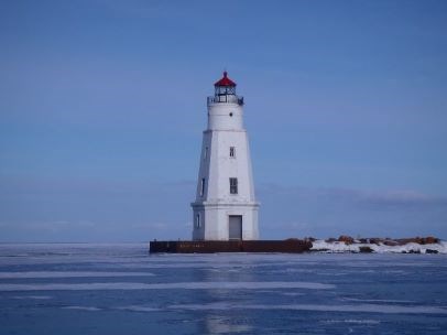A white lighthouse with a red roof stands on a break wall on a frozen lake.