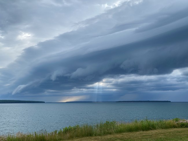 Dark clouds show a storm front moving over a large lake with a small forested island in the distance.
