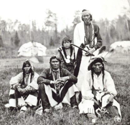 A historic photo of five Ojibwe men in traditional clothing, gather in front of a couple round bark covered lodges.