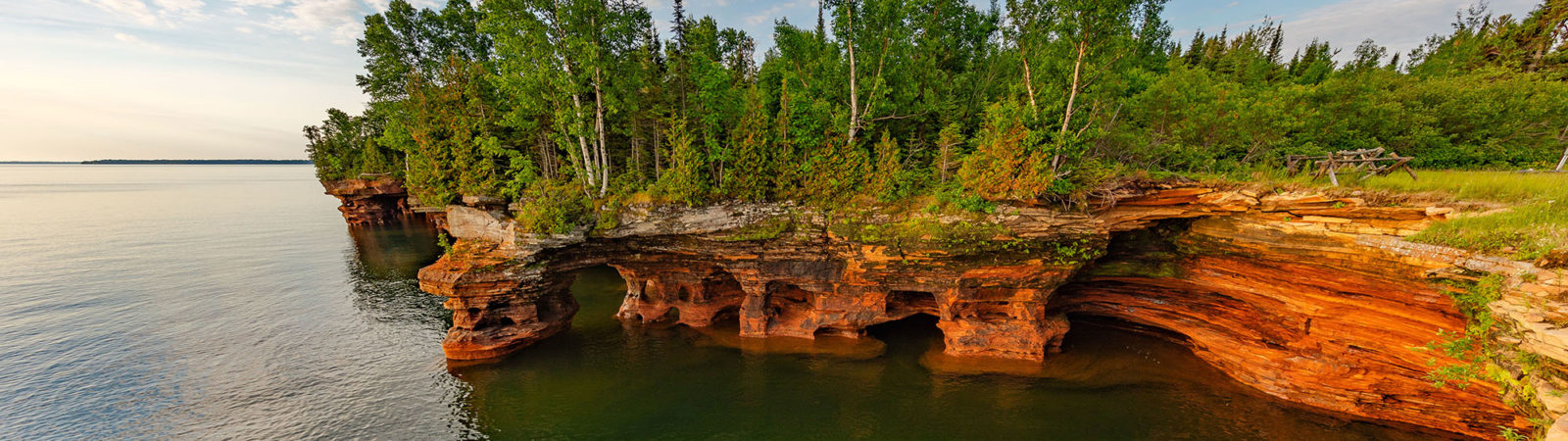 Tree covered red cliffs, carved with caves, meet the blue water.