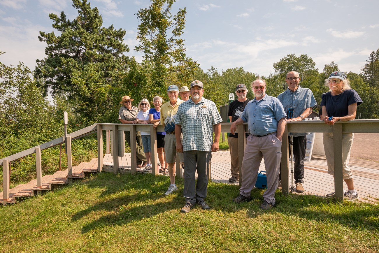 Photograph of group of people looking towards camera with a staircase to their right, trees in the background, and a blue sky.