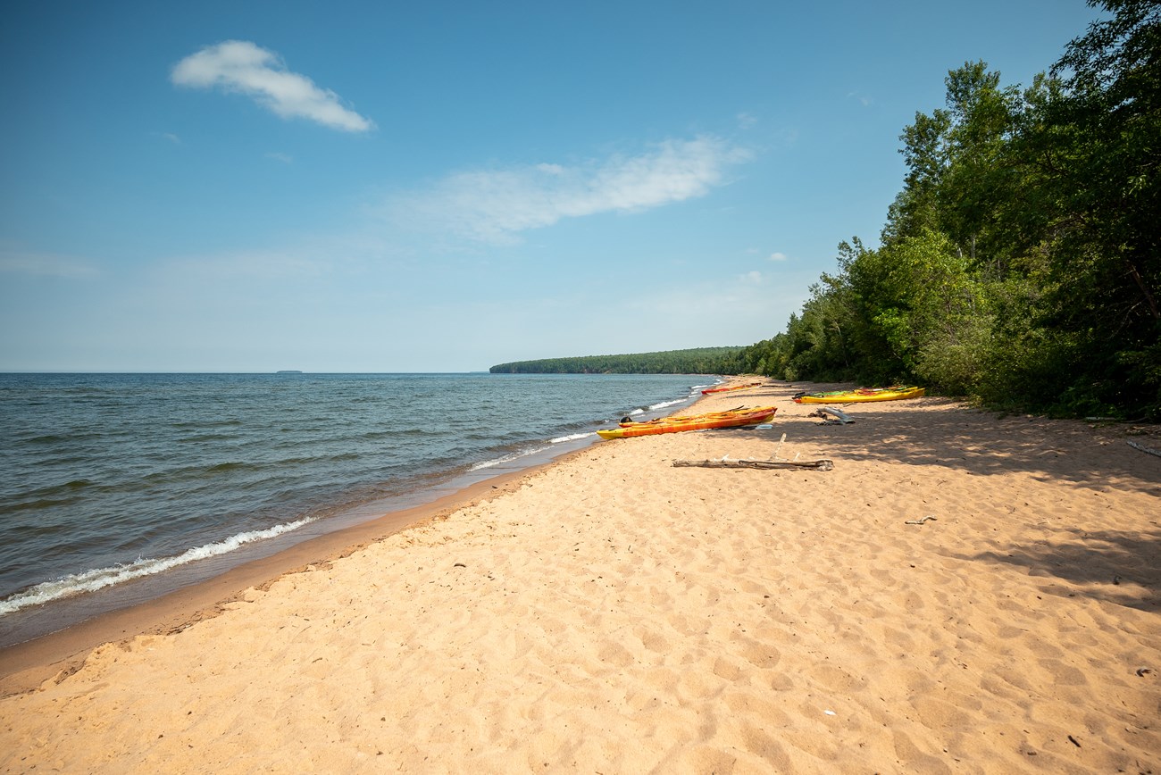 Meyers Beach Photograph of a long sandy beach with Lake Superior on the left and trees on the right. Colorful orange and yellow kayaks line the sand.