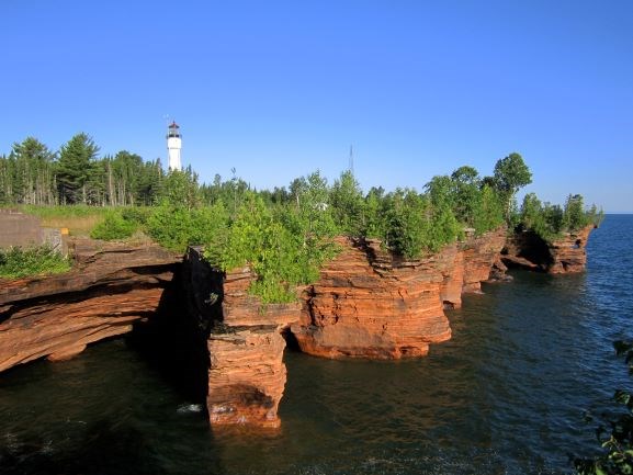 Sandstone cliffs on lake shore with trees and white lighthouse tower on top of cliffs.