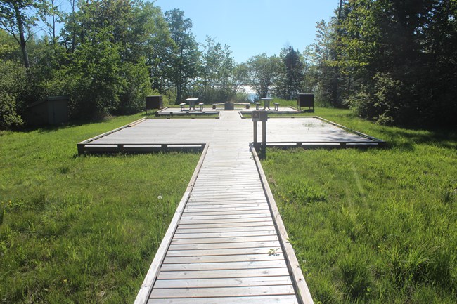 A boardwalk leads to a larger platform with a picnic table, fire ring, and bear-proof storage locker.