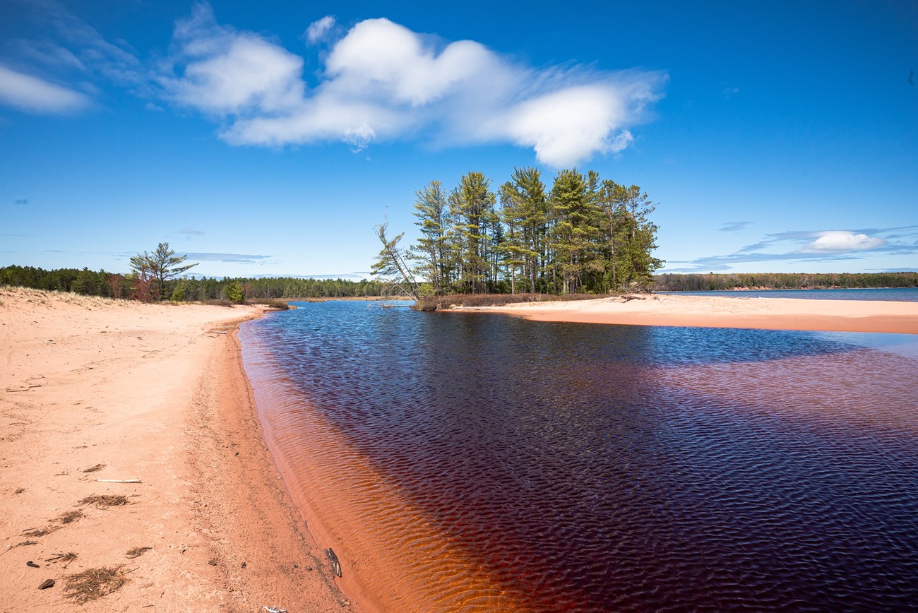 Photograph of sandy beach, dark lake water, and trees under a partly cloudy blue sky.