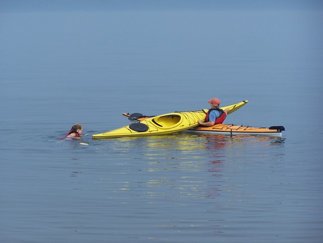 A person in a kayak assists another person in the water with the front of their kayak on top of their own.