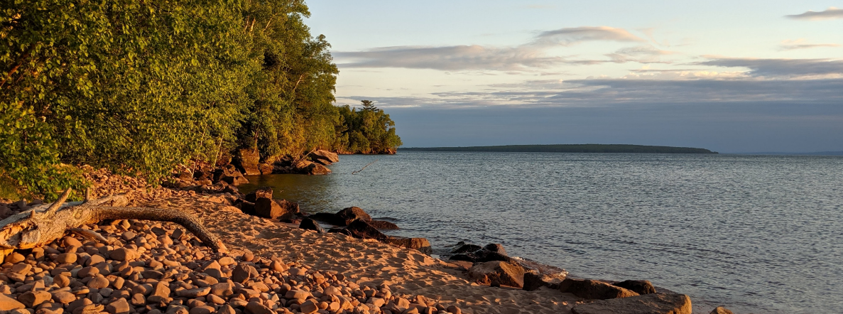 Evening sun light hits the rocky shore with a lake and forested island in the distance.