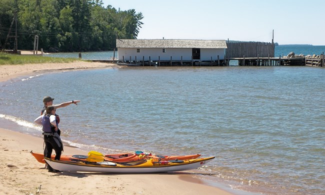 Two people standing on a beach next to kayaks pointing out at the lake.