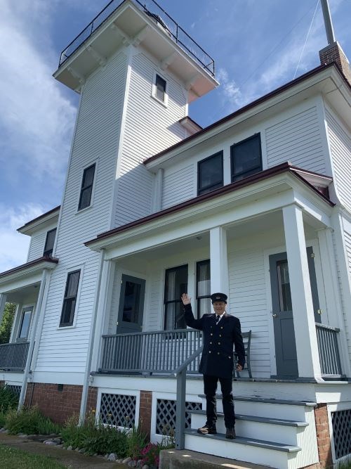 A Park Ranger dressed in a dark suit and hat as a lighthouse keeper waves from the front porch of a white house with red trim.