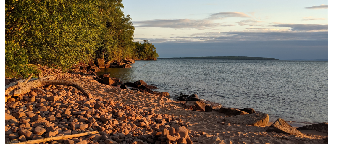 The glow of a cobble beach near the edge of a forest looks out to a forested island in the distance at sunset.