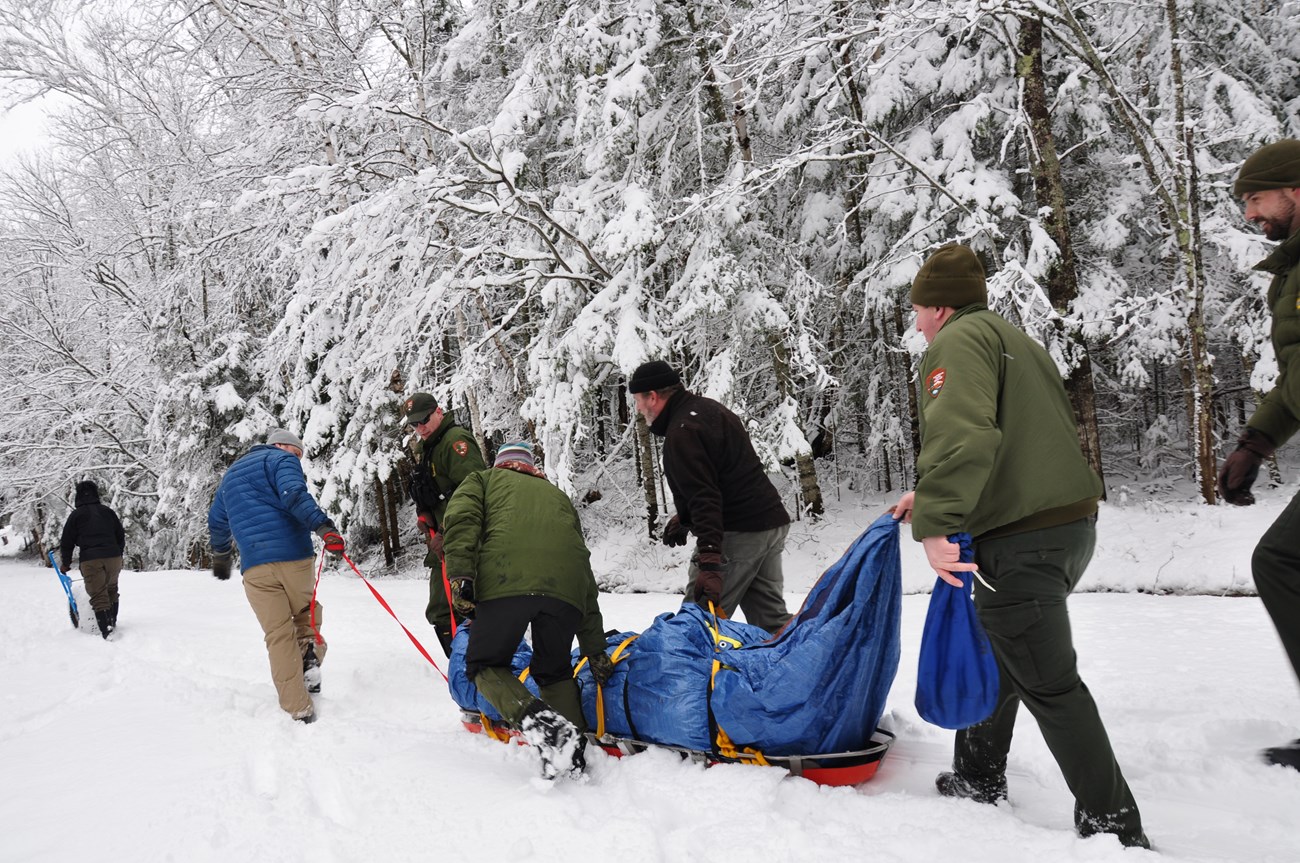 6 park rangers pull a patient wrapped in a tarp on a sled in the snow.