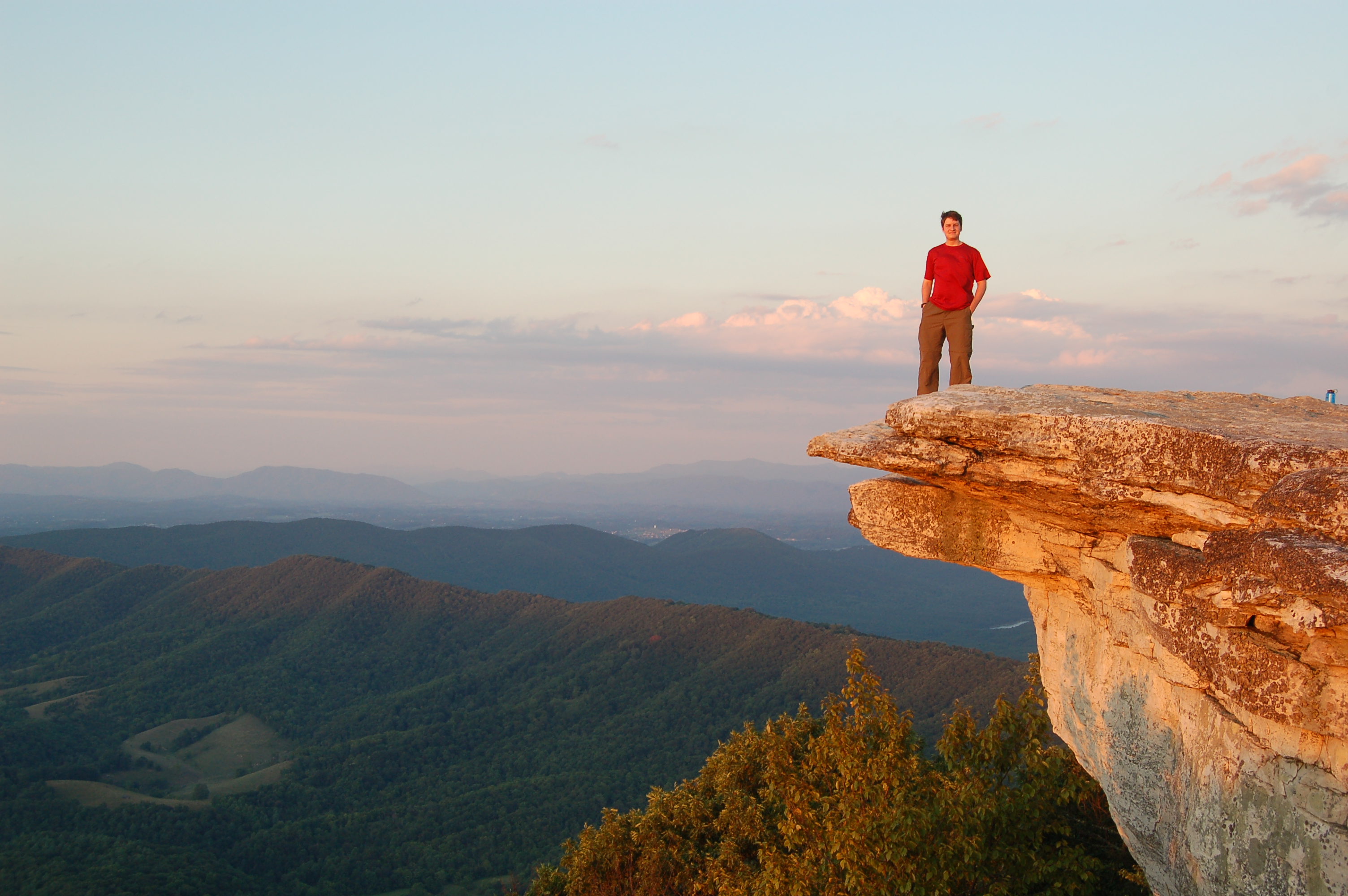 A man happily stands near the edge of a very steep cliff overhang View of man standing on overlook along the Appalachian Trail in the Triple Crown section.