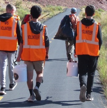 Volunteers walk down a paved path, wearing orange vests that say "NPS VOLUNTEER" while carrying buckets and trash pickers.
