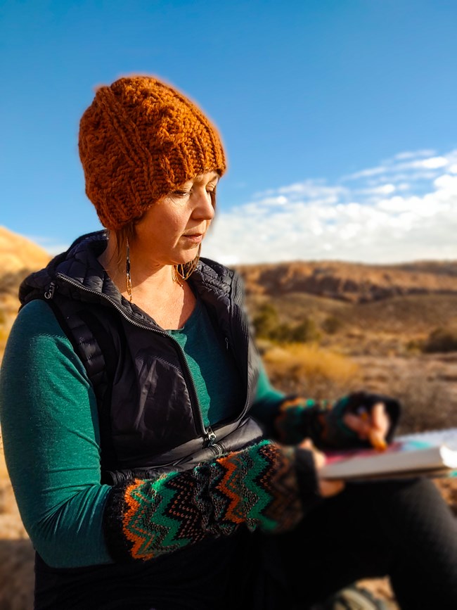 light-skinned woman in orange knit hat sketches in notebook; tan rocky backdrop and blue sky
