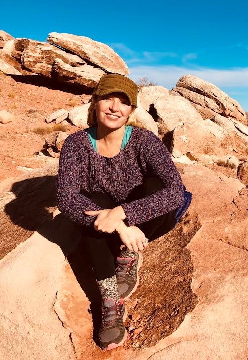 A woman wearing a purple sweater, brown hat, and hiking shoes, seated on orange rocky slope with a blue sky in the background.