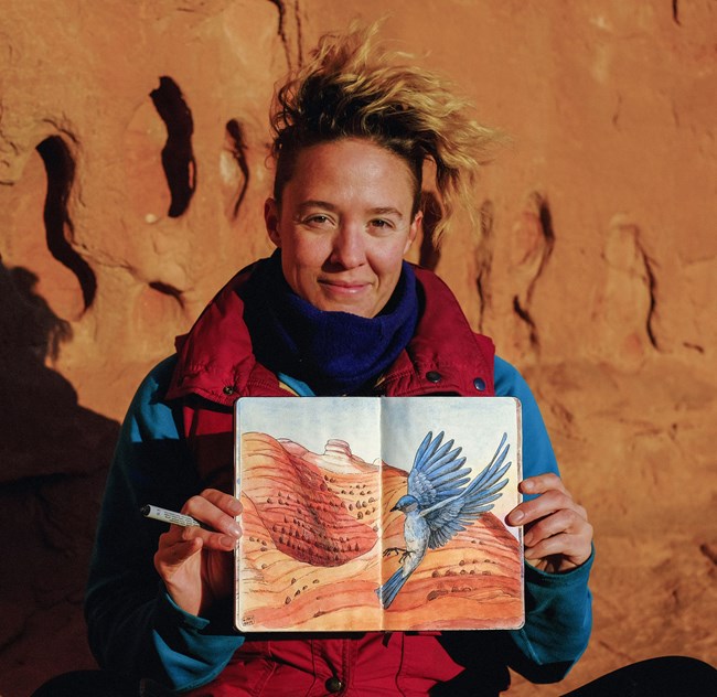 A woman holding up a sketchbook showing an illustration of a blue bird flying in front of a red rock landscape. The woman is sitting outside, in front of a red rock formation, wearing a blue and red jacket.