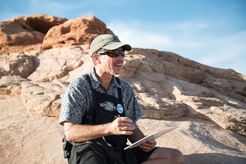 a man holds a paint brush and canvas in front of a rock outcrop