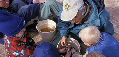 A volunteer and children look over a bucket filled with soil A volunteer and children look over a bucket filled with soil