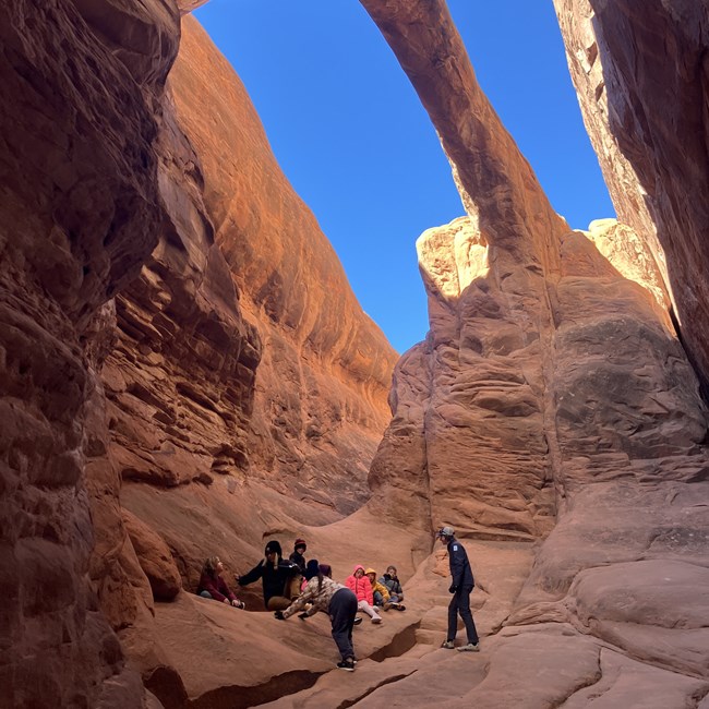 Group of children with one adult standing underneath a red sandstone arch. Canyon walls surround the group. Blue sky is above.
