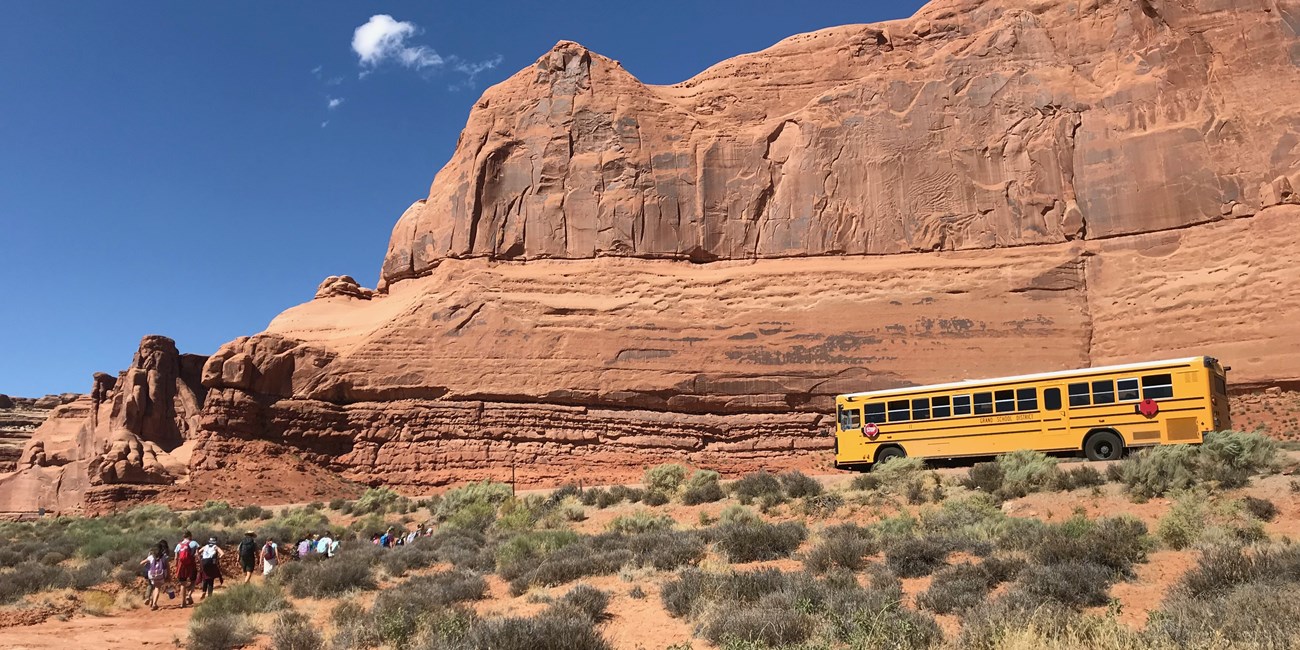 A school bus is parked along a road near a large red sandstone wall. A group of students with a park ranger hike on a path through shrubs and grasses towards the bus.