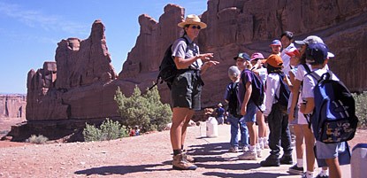 A park ranger on a trail, wearing shorts and a flat hat, talking to a group of second graders. Eroded red rock fins are in the background with a bright blue sky.