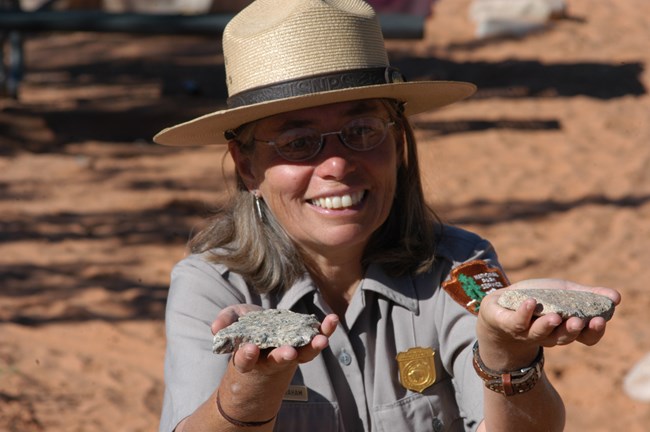 A woman wearing a park ranger uniform and straw hat holds two gray rocks up near her face.