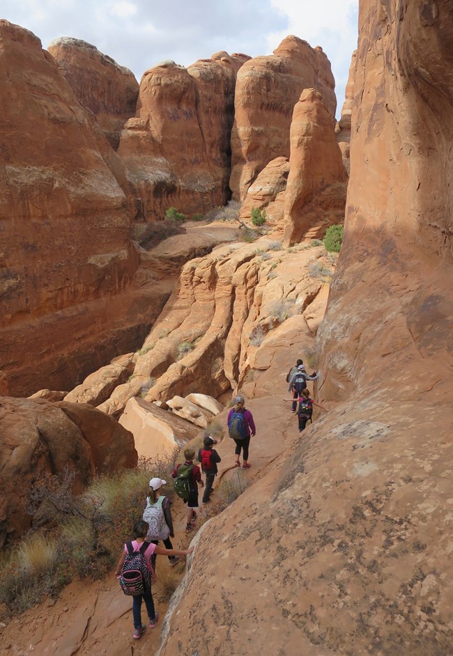A group of children hike along a narrow section of sandstone between canyon walls.