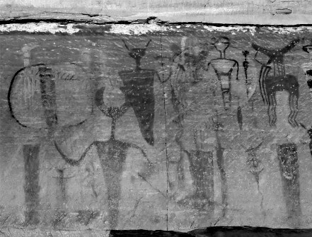 Black and white image depicting rock art on surface. Image shows several dark humanoid figures on a lighter background. The image also features several animal like figures with antlers or horns.