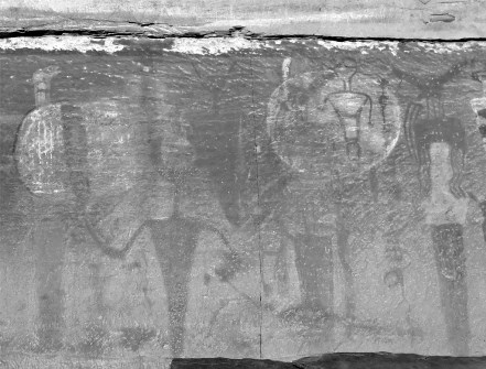 A close up black and white image of rock art. Rock art shows several humanoid figures and two spheres with dark lines running vertically through them.