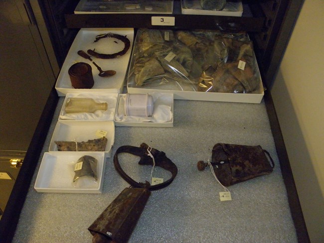 Photograph of contents in a museum cabinet. A historic cow bell and sheep bell, glass containers, a horseshoe, a fork and a tin can.