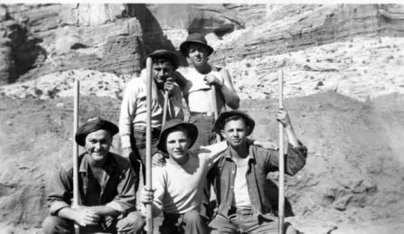 A black and white image of five young men posing for a photo while holding shovels in front of a rockface.