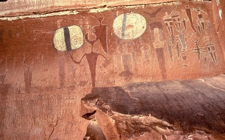 Image of rock art on red sandstone. The drawings feature more than a dozen humanoid figures in shades of brown, red, and white.
