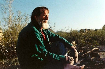 A dark haired man with a white beard sits cross legged and barefoot among green shrubs. He looks off in the distance.