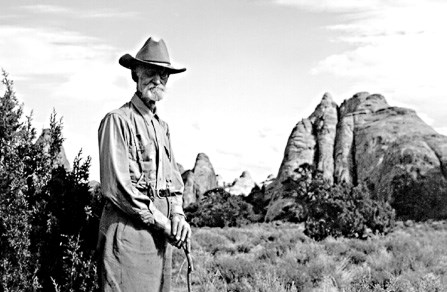 A black and white photo man wearing a cowboy hat stands among shrubs leaning on a walking stick. A row of large rock fins can be seen in the background.