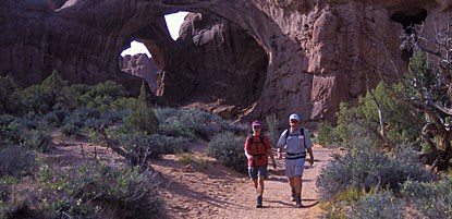 A pair of hikers walk on a sandy trail with large sandstone arches in the background