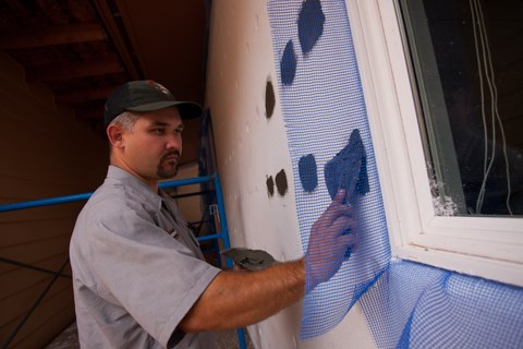 A worker stand next to scaffolding applying putty to a wall with a putty knife.