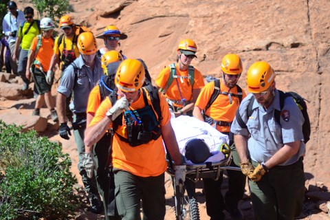 a line of people wearing bright orange helmets and shirts carrying a person in a wheeled litter down a red sandstone trail.