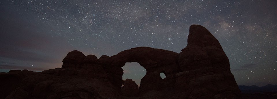 A bright starry sky behind the silhouette of a dark rock wall containing two arches.