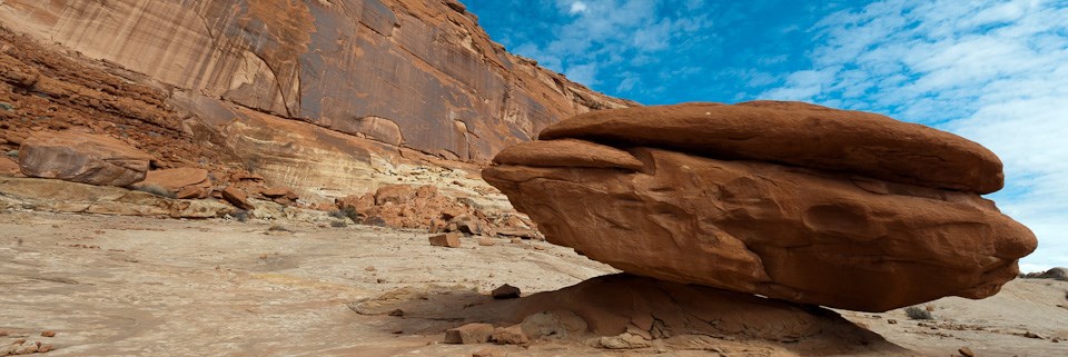 a broad reddish orange, flat rock, balanced on a smaller rocky shelf. Background has high red colored cliff face and a blue sky.